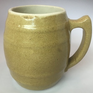 May include: A light brown ceramic mug with a rounded body and a handle. The mug has a slightly textured surface.