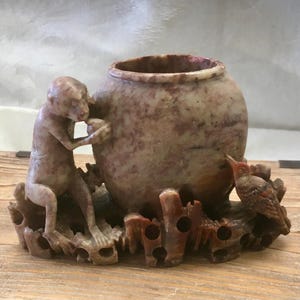 May include: A carved stone vessel featuring a mottled reddish-brown finish. The container is adorned with a sculpted monkey figure on the left side and a small bird perched on the right, all resting on a base with openwork, branch-like detailing.