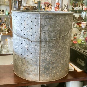 May include: A large, cylindrical galvanized metal bucket featuring a uniform pattern of small, evenly spaced circular perforations throughout its body. The bucket includes a sturdy, arched wire handle attached to the sides for carrying.