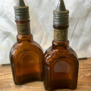 May include: Two vintage brown glass bottles with metal stoppers. The bottles have a unique shape and a label that reads "Stetson". The bottles are displayed on a wooden surface.