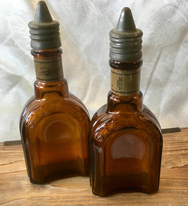 May include: Two vintage brown glass bottles with metal stoppers. The bottles have a unique shape and a label that reads "Stetson". The bottles are displayed on a wooden surface.