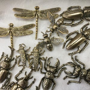 May include: A collection of antique brass-colored insect figurines, including dragonflies, bees, and beetles. The detailed metalwork showcases the wings and body segments of each insect. The figurines are arranged on a white surface.