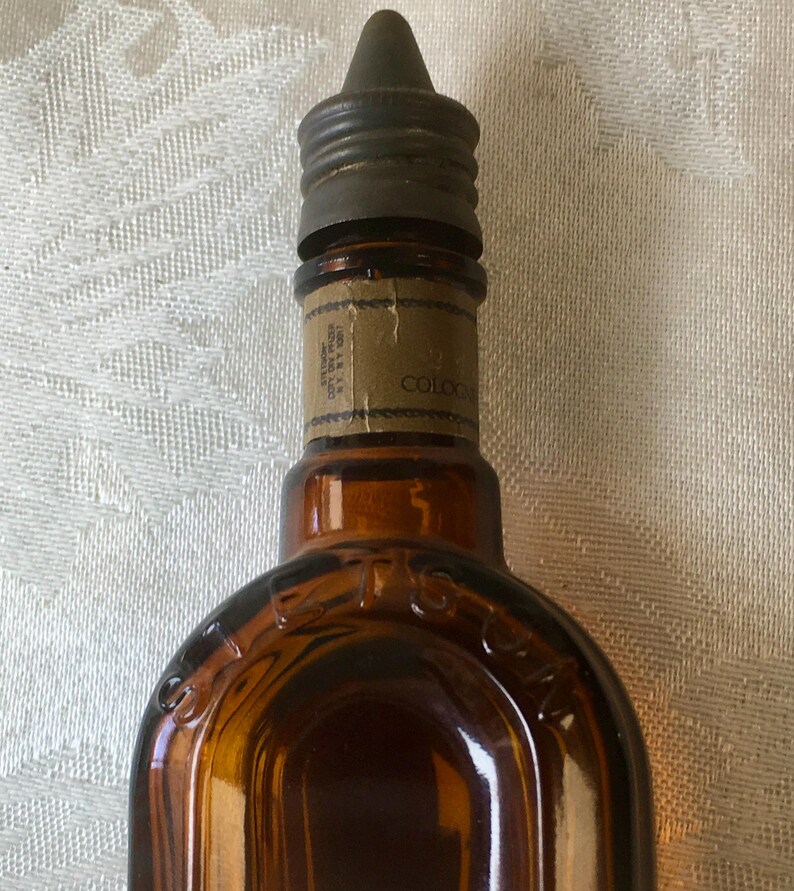 May include: Close-up of a vintage brown glass cologne bottle with a metal cap. The bottle has a label with the word "COLOGNE" visible. The bottle's shape is rounded at the bottom and tapers towards the top.