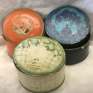 May include: Three round metal tins with lids. One is orange with a circular illustration of two figures. One is dark blue with a silhouette of a person under a tree. One is cream with a green grid pattern, floral accents, and text reading Poudre de Toilette April Showers Cheramy Paris New York.