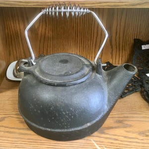 May include: A black cast iron teapot with a silver metal handle and a coiled spring grip. The teapot has a round body, a lid, and a spout. It sits on a wooden shelf.