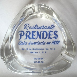 May include: A clear glass ashtray with a blue and white logo that reads "Restaurante PRENDES Casa fundada en 1892" with the address "Av. 16 de Septiembre No. 10-C México 1. D. F. 512-09-94 513-06-16".