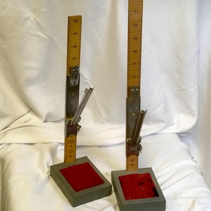 May include: Two vintage wooden measuring tools with metal components. Each tool features a ruler marked in inches, attached to a metal mechanism and set on a gray base with a red velvet insert. The rulers measure up to 21 inches.