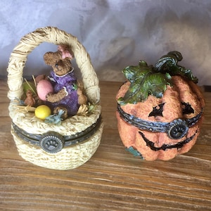 May include: Two decorative trinket boxes. One is a basket with a bear figurine holding an egg. The other is an orange pumpkin with a carved face and green leaves. Both boxes have hinged lids and are on a wooden surface.