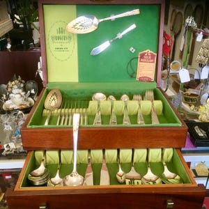 May include: A wooden silverware chest with a green velvet interior, filled with silver utensils. The chest is open, revealing multiple drawers and compartments. The top of the chest has a green background with two silver serving utensils. The chest is labeled "1881 Rogers Silverplate".