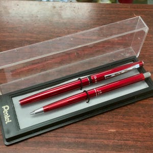 May include: A set of two metallic red Pentel writing instruments, a ballpoint pen and a mechanical pencil, housed in a clear plastic display case with a black base. The brand name Pentel is printed in white on the left side of the black case base.