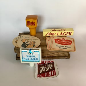 May include: A collection of vintage beer-related items, including a Piels tap handle, Yuengling Light LAGER sign, coasters, and a Schlitz Malt Liquor tap and patch. The items are arranged on a wooden surface.