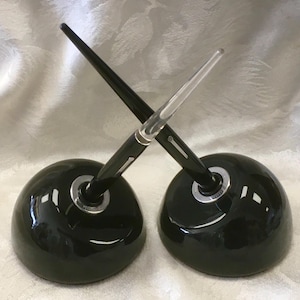 May include: Two black pen holders with pens. Each holder is a glossy, dark green sphere with a silver ring. The pens are black with clear tips, crossing each other. The background is a light, textured fabric.