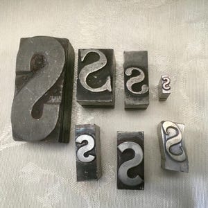 May include: A collection of vintage metal letterpress printing blocks, each featuring the letter "S". The blocks vary in size, with a large, rectangular block dominating the composition. The metal has a worn, aged appearance.