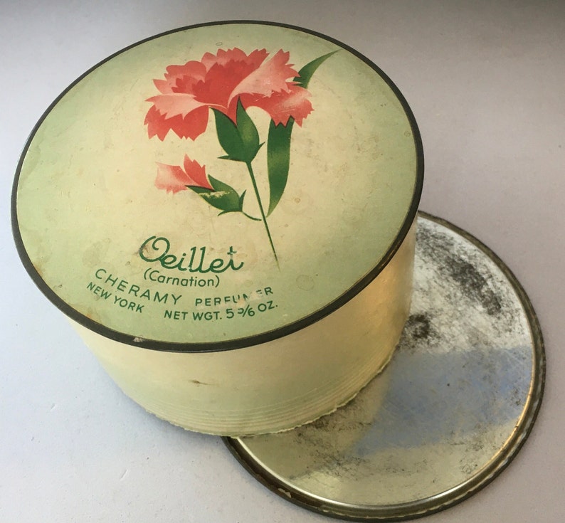 May include: A vintage round powder compact with a green and white lid. The lid features a pink carnation flower with green leaves and the text "Oeillet (Carnation) CHERAMY PERFUMER NEW YORK NET WGT. 5 3/8 OZ."
