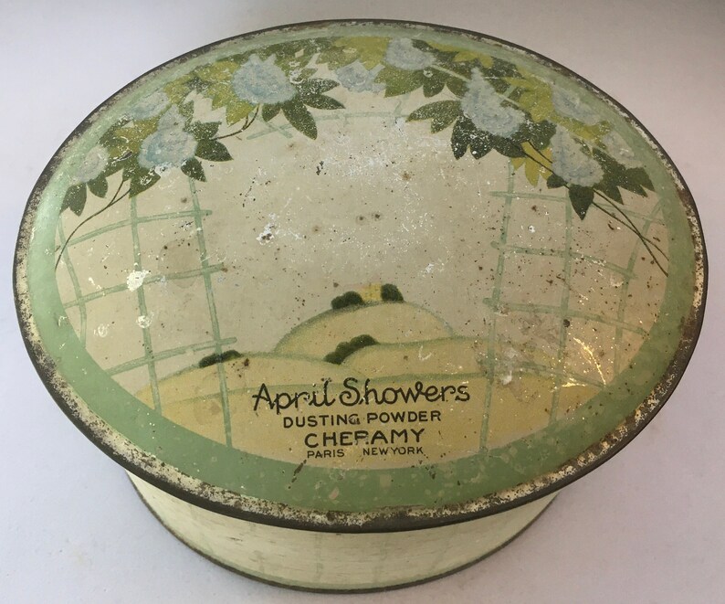 May include: Vintage round tin container with a green and white floral design. The lid features a green and white landscape with the text "April Showers Dusting Powder  Cheramy Paris New York".