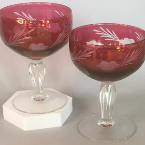 May include: Two ruby red stemmed glasses with etched white floral designs. The glasses are on a white hexagonal base.
