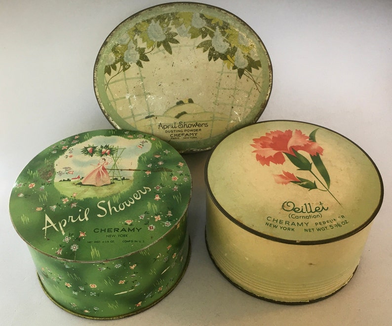 May include: Three vintage round powder tins with floral designs. The top tin is green with white flowers and the text "April Showers Dusting Powder Cheramy New York". The middle tin is green with white flowers and the text "April Showers Cheramy New York Net Wt 4 1/16 oz Comp in U.S.". The bottom tin is green with a pink carnation and the text "Oeillet (Carnation) Cheramy Persua New York Net Wt 5 3/4 oz."