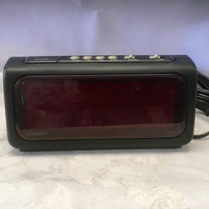 May include: Black Cosmo digital alarm clock with a red LED display. The clock has buttons for snooze, hour, minute, time, alarm, and dimmer. The word "Cosmo" is printed on the front.
