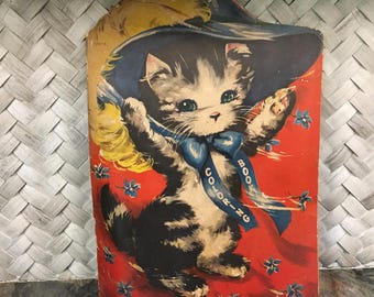 Puede incluir: Libro para colorear vintage con una ilustración de un gatito con sombrero y lazo azules, sobre un fondo rojo con motivos florales azules. El gatito sostiene una pluma amarilla y una cinta azul con la inscripción "Coloring Book".