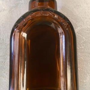 May include: A vintage brown glass bottle with a rounded top and a label on the neck. The bottle has a rectangular shape with rounded edges and is filled with a dark liquid. The word "STETSON" is embossed on the top.