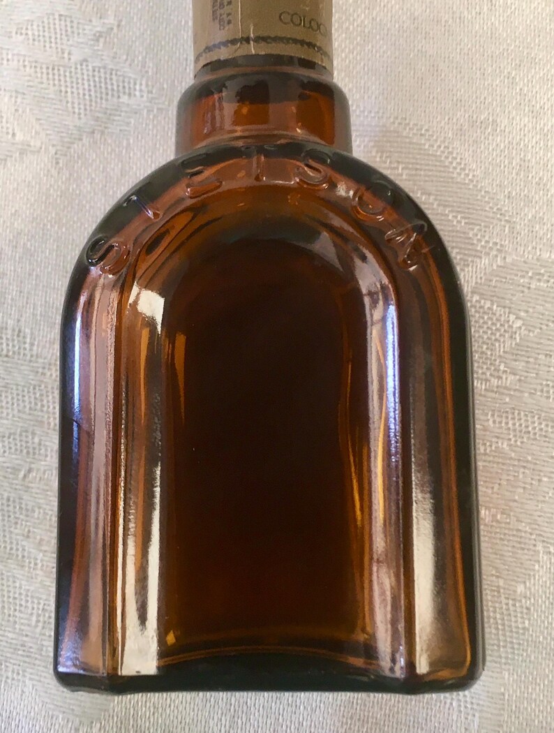 May include: A vintage brown glass bottle with a rounded top and a label on the neck. The bottle has a rectangular shape with rounded edges and is filled with a dark liquid. The word "STETSON" is embossed on the top.