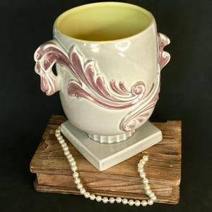 May include: A gray ceramic planter with a square base and two handles. The planter has a raised design of pink and gray leaves and swirls. A string of white pearls is in front of the planter.