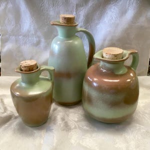 May include: Three ceramic pitchers with handles and cork stoppers. Each vessel features a smooth, glazed finish with a gradient color scheme transitioning from a soft mint green at the top to a warm, earthy brown tone at the base of the containers.