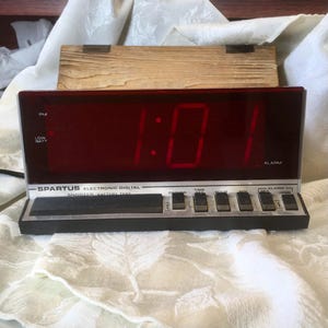 May include: A vintage Spartus electronic digital alarm clock with a red LED display showing 1:01. The clock has a black and silver base with buttons for time setting and alarm functions. A wooden box is behind the clock.