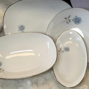 May include: A set of four white porcelain serving dishes with a silver trim and a blue rose design. The set includes a large oval platter, a round plate, and two smaller oval dishes. The dishes are arranged on a white surface.