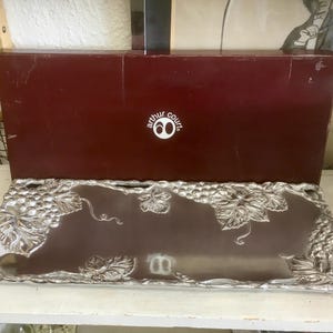 May include: A rectangular silver-toned metal serving tray featuring a raised grape and vine motif along the edges. The tray is positioned in front of a dark maroon rectangular box with the white text "arthur court" and a "60" logo printed on it.