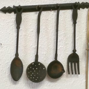 May include: A set of four antique-style kitchen utensils hanging on a decorative wrought iron bar. The set includes a spoon, a slotted spoon, a ladle, and a fork, all in a dark, aged finish.