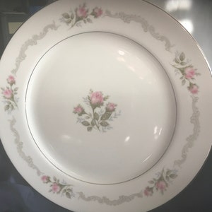 May include: White ceramic dinner plate with a gold rim and a floral design featuring pink roses and gray leaves. The roses are clustered in the center of the plate and are repeated around the rim.