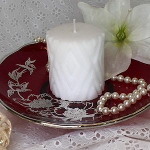 May include: A white pillar candle with a textured design sits on a red plate with silver floral accents. A string of white pearls and a white flower are in the background.