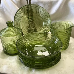 May include: A collection of green glass serving dishes, including a bowl, plates, a pitcher, and a covered jar. The glass has a textured, ridged pattern. The set is arranged on a white surface.