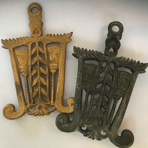 May include: Two decorative metal trivets, one gold and one black, with a design of three stylized leaves and a central stem. Both trivets have a scalloped edge.