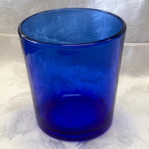May include: A cylindrical drinking glass made of deep cobalt blue glass. The vessel features a smooth, uniform exterior surface with a thick base and a simple, open rim design, showcasing a vibrant and saturated blue color throughout the piece.