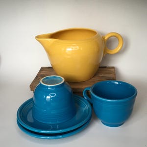 May include: A yellow ceramic pitcher with a handle and spout, alongside a set of turquoise-colored ceramic teacups and saucers. The teacups and saucers are stacked and arranged on a wooden surface, creating a colorful kitchenware display.