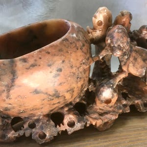 May include: A carved stone vessel featuring a smooth, rounded bowl attached to a base with intricate, openwork detailing. The stone is a mottled mix of peach, tan, and dark brown hues, with sculpted figures integrated into the side of the bowl.