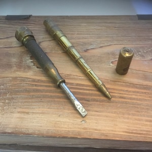 May include: Two brass tools on a wooden surface. One tool is a screwdriver with a brass handle and a metal blade. The other tool is a telescoping pen with a brass body and a metal tip.