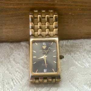 May include: A rectangular gold-tone wristwatch with a black dial, featuring a crown logo, gold-tone hands, and two small clear stones at the twelve and six o'clock positions. The watch has a textured gold-tone metal link bracelet.
