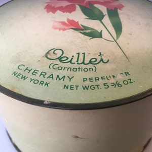 May include: A vintage round powder compact with a green and white label. The label features a pink carnation with green leaves and the text "Oeillet (Carnation)" in cursive. The text "CHERAMY PERFUMER NEW YORK NET WGT. 5 3/6 OZ." is also on the label.