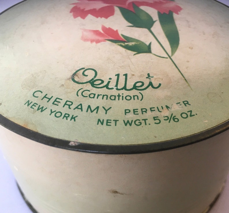 May include: A vintage round powder compact with a green and white label. The label features a pink carnation with green leaves and the text "Oeillet (Carnation)" in cursive. The text "CHERAMY PERFUMER NEW YORK NET WGT. 5 3/6 OZ." is also on the label.
