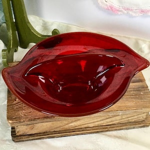May include: A vintage, ruby red glass dish with a unique, organic shape. The dish has a shallow, oval bowl with gently curved sides and a slightly raised rim. It sits on a wooden surface, showcasing its rich color and design.