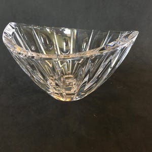May include: A clear, decorative glass bowl with a unique, angular design. The bowl has a wide, open top and a series of vertical, textured lines that create a geometric pattern. The bowl is made of clear glass, allowing light to pass through.