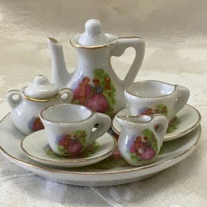 May include: A miniature porcelain tea set with a white base and gold trim. The set includes a teapot, sugar bowl, creamer, two teacups, and saucers. Each piece features a painted scene of figures in a garden setting.
