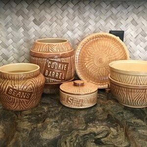 May include: A set of five brown ceramic kitchen canisters with wood grain patterns. The canisters are labeled "Cookie Barrel", "Beans", and "Save Store Serve".