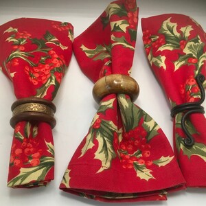 May include: Three red fabric napkins with a holly and berry pattern are folded and held together with different napkin rings. One ring is made of wood, one is made of metal, and one is a simple, ornate metal ring.