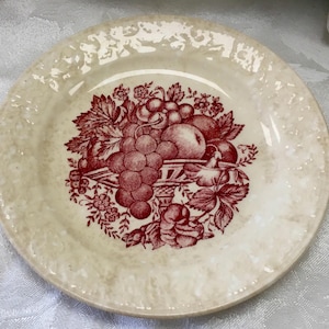 May include: A round, cream-colored plate with a red fruit basket design in the center. The design includes grapes, apples, and other fruits. The plate has a textured rim.