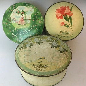 May include: Three vintage round powder compact tins. Two are green with white floral designs and the text "April Showers". One is white with a red carnation and the text "Oeillet (Carnation) CHERAMY PERFUMS R NEW YORK NET WT. 5/8 OZ."