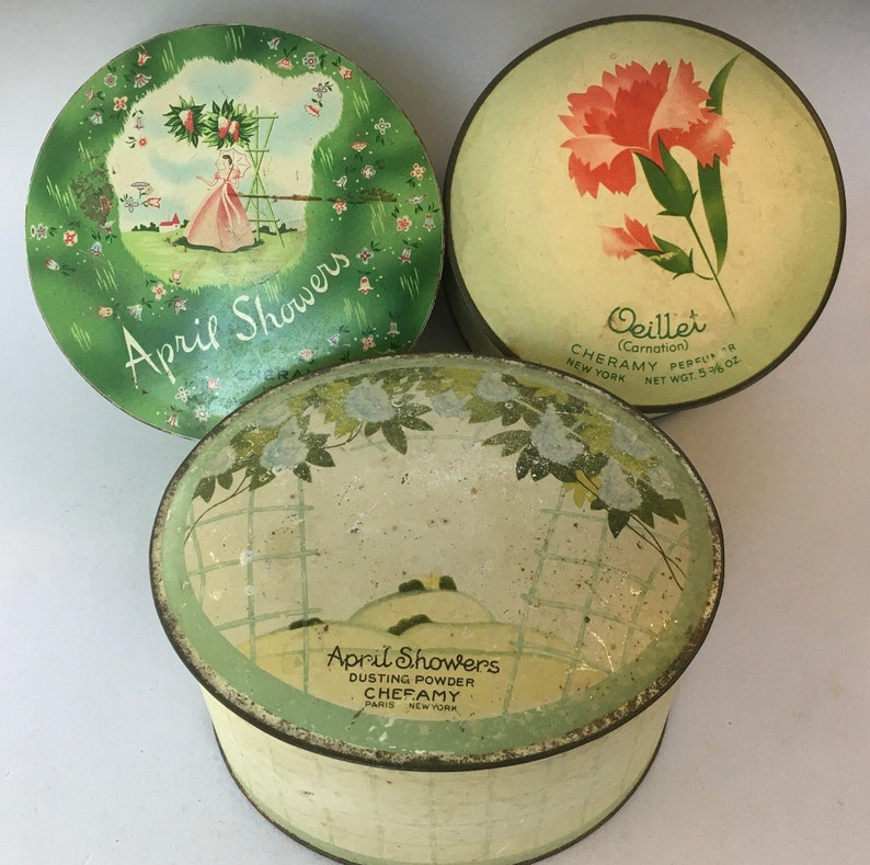 May include: Three vintage round powder compact tins. Two are green with white floral designs and the text "April Showers". One is white with a red carnation and the text "Oeillet (Carnation) CHERAMY PERFUMS R NEW YORK NET WT. 5/8 OZ."